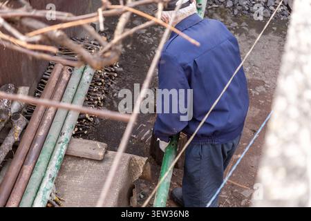 An elderly person in a blue jacket performs outdoor chores by moving metal poles, capturing the essence of active aging, home improvement, and manual Stock Photo