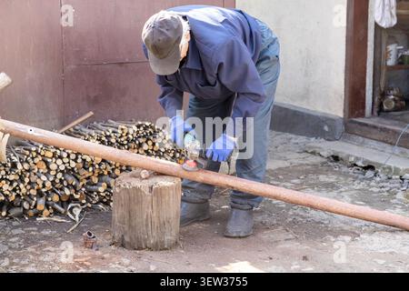 A person wearing protective blue gloves uses an angle grinder on a long corroded iron pipe placed on a tree stump, illustrating construction repair wo Stock Photo