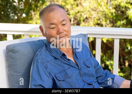 Mature Asian man sitting on chair with blue-gray cushion on white deck railing, wearing denim shirt Stock Photo