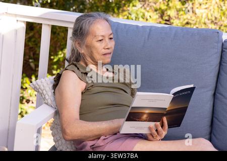 Senior Asian woman reclining on blue-gray porch sofa, leaning on pillow, reading paperback Stock Photo