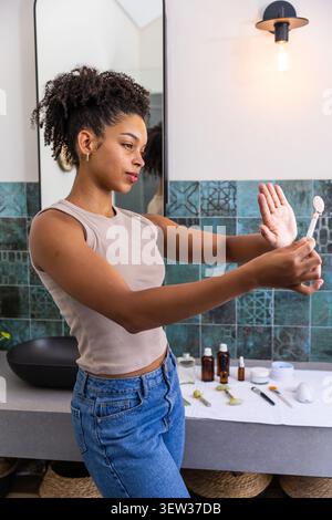 African American woman examining face using handheld mirror at vanity, wearing sleeveless beige top Stock Photo