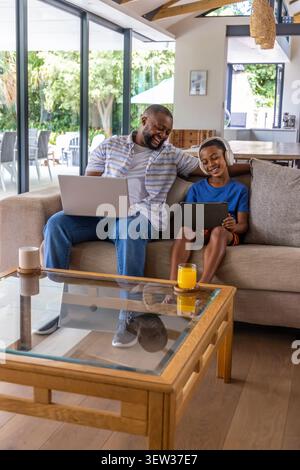 African American man and boy sitting on beige sofa sharing tablet, laptop, headphones, orange juice Stock Photo