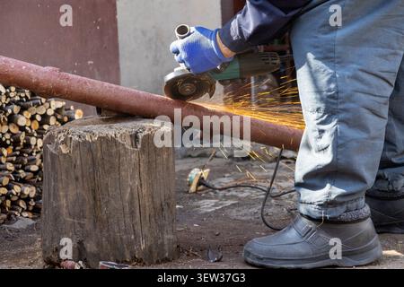 A person in protective gear cuts iron tubing with a power tool, creating bright sparks in a gritty workshop to demonstrate professional construction s Stock Photo