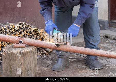 A professional handyman uses an angle grinder to cut through an oxidized iron pipe in an outdoor workshop, highlighting precision metalwork, manual la Stock Photo