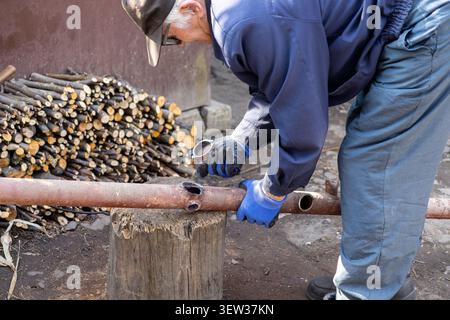 Mature craftsman working outdoors with a corroded iron pipe, using a log as a makeshift workbench to convey skilled labor, manual effort, and practica Stock Photo