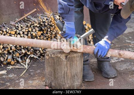 A person cuts an old metal pipe with an electric power tool on a log, capturing the intensive manual labor, flying sparks, and precise craftsmanship i Stock Photo