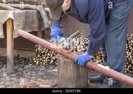 A focused craftsman wearing protective gloves operates a hand-held power grinder on a steel tube at a busy lumber yard, highlighting precision, hard w Stock Photo