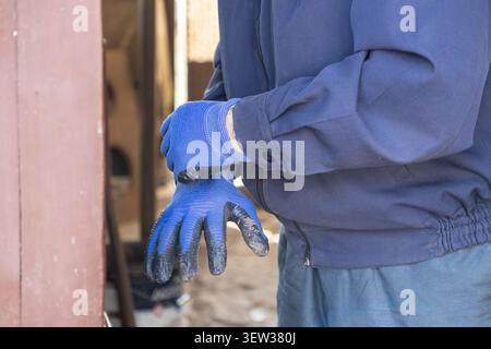 Person wearing protective gear pulls on blue utility gloves to start physical labor at an industrial site, illustrating focus on task preparation and Stock Photo