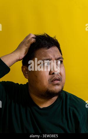Confused bearded Asian male scratching his head while looking to the side with a skeptical expression against a solid bright yellow background. His fi Stock Photo