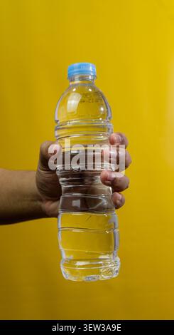 A close view of a hand firmly holding a transparent plastic bottle filled with fresh water against a vibrant yellow background. The clear bottle featu Stock Photo