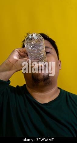 An adult male wearing a dark green shirt drinks fresh water directly from a transparent plastic bottle against a vibrant solid yellow background. The Stock Photo