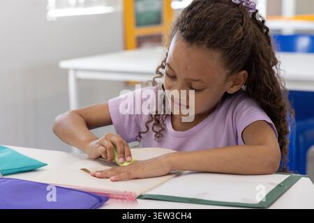 African American child student erasing in notebook using eraser at desk in class light purple shirt. Classroom, daylight, focus, pencils, pouches, scrunchie, folder Stock Photo