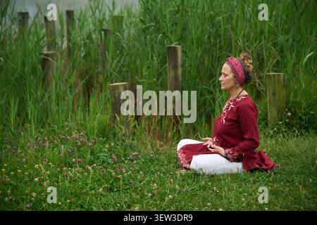 Woman in traditional attire practicing meditation outdoors, sitting cross legged on a grassy field surrounded by lush green reeds, promoting mental cl Stock Photo