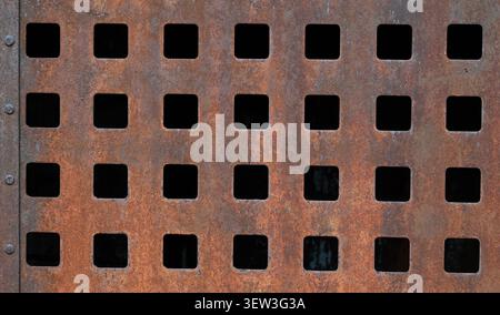 Close up of rusted metal surface with pitted corrosion, cutout grid pattern, dark openings, and vertical row of rivets Stock Photo