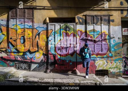 A man wearing a blue beanie and puffer jacket leans casually against a richly textured, graffiti-covered building wall in Istanbul, Turkey, engrossed Stock Photo
