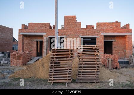 A construction site showing the ongoing building process of a residential house made of red bricks, with stacks of wooden pallets and piles Stock Photo