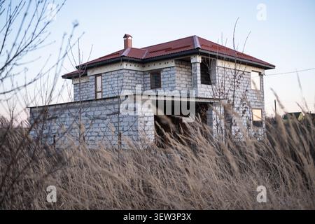 A view of an unfinished two-story house built from cinder blocks with a red metal roof, framed by dry, tall autumn grass in the foreground on a calm Stock Photo