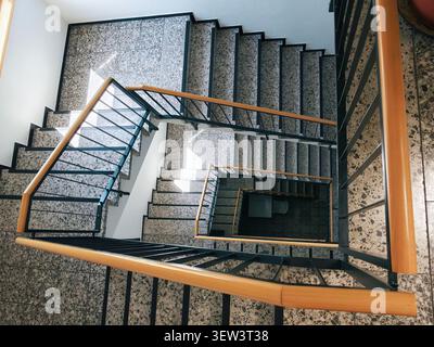 Top down view of multi level indoor staircase with terrazzo steps, black metal balustrades, and smooth wooden handrails, lit by natural window light, Stock Photo