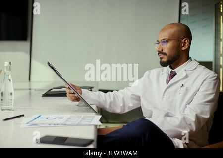 Middle aged man doctor using tablet while reviewing medical data at desk, supporting digital healthcare workflow. Useful for telemedicine, clinic management, patient records marketing Stock Photo