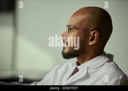 Close up of South Asian male doctor wearing glasses, looking away and reflecting during workday, conveying healthcare expertise and decision making. Ideal for medical branding Stock Photo