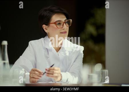 Medium shot of middle aged Caucasian woman doctor sitting with pen, listening during consultation or meeting. Suitable for healthcare communication, clinic marketing, expert advice Stock Photo