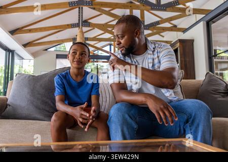 Father and adolescent son sitting on beige sofa in living room, father pointing and talking Stock Photo