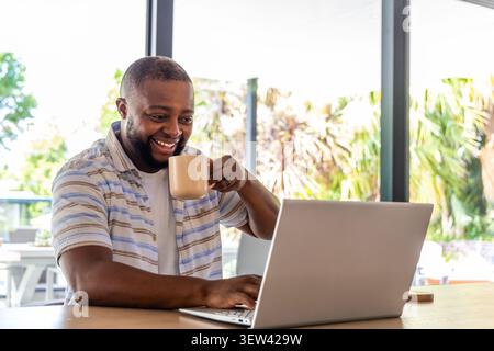 Man wearing striped shirt sitting by large windows, using silver laptop and holding ceramic mug Stock Photo
