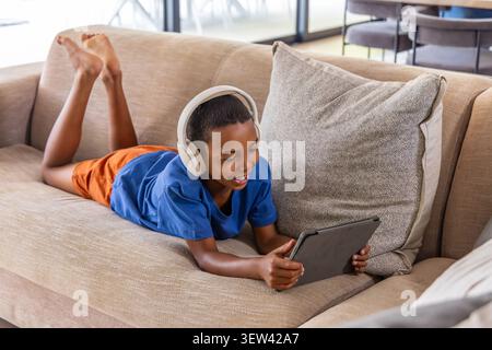 School-age male child lying on beige sofa in living room holding tablet wearing white headphones Stock Photo