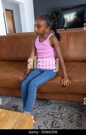 African American youth wearing pink top and jeans sitting on leather couch at home, looking left Stock Photo