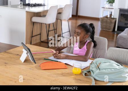 African female child sitting at dining table using tablet on stand, writing in notebook, copy space Stock Photo