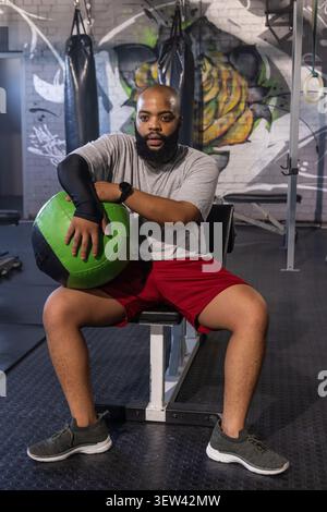 African American man in sportswear sitting on bench in gym holding green black medicine ball Stock Photo