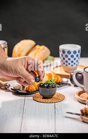 In the tranquil atmosphere of a morning, bathed in natural light, a hand carefully places blueberries, one by one, into a small, handcrafted ceramic b Stock Photo