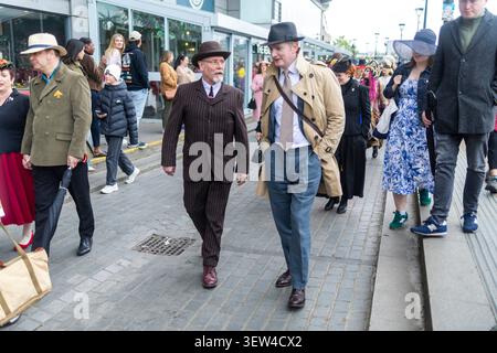 London, UK, 10 May 2026, London Hat Walk gentlemen in classic vintage suits, coats, and fedora hats walking and conversing on a paved city sidewalk du Stock Photo