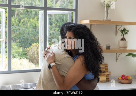 Diverse couple hugging in bright kitchen by multi-pane window, woman showing smartwatch, fruit bowl Stock Photo