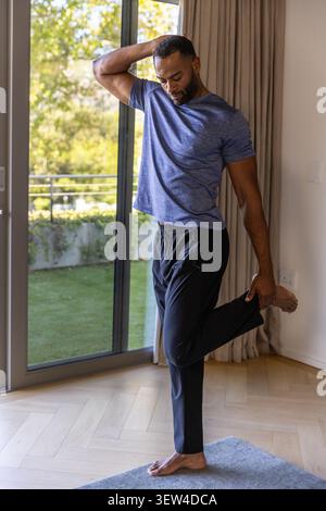 African American senior man stretching quad on gray rug by glass door barefoot wearing blue shirt Stock Photo