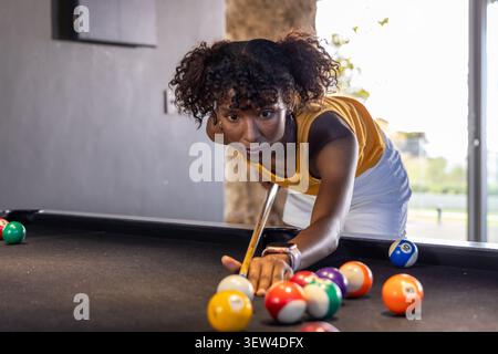 Woman aiming shot on black-felt pool table with cue stick and cue ball, wearing yellow tank Stock Photo