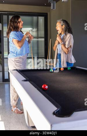 Diverse female friends standing holding wood cues talking by black pool table in home game room Stock Photo