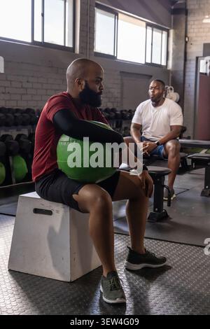 African men sitting on plyometric box holding green medicine ball at gym, wearing workout clothes Stock Photo