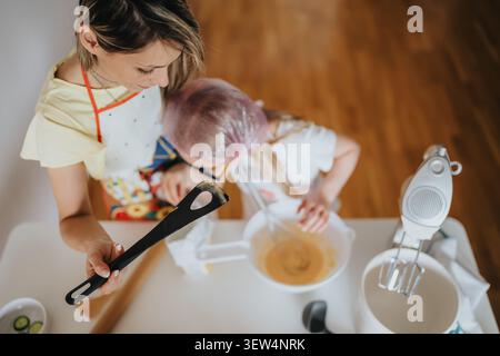 Mom and child baking together at home mixing batter in a bright kitchen while wearing aprons Stock Photo