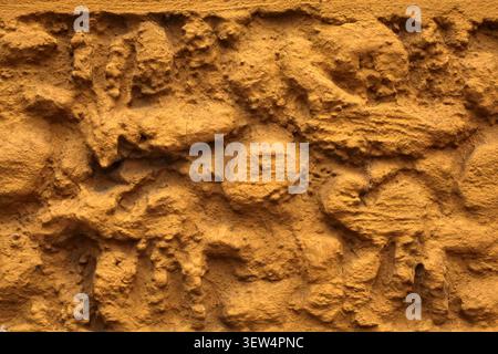 A full-frame close-up capturing the extremely rough, uneven, and heavily textured surface of a plaster stucco wall painted in mustard yellow. Stock Photo