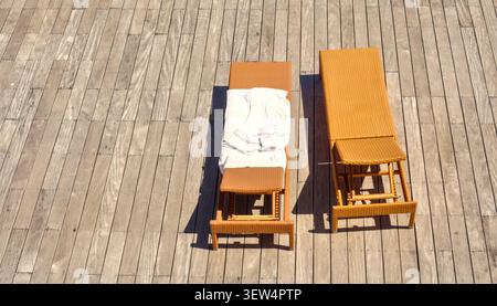A high-angle, top-down view of two empty brown wicker sun loungers placed on a light-colored wooden plank deck, one with a white towel. Stock Photo