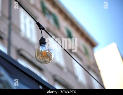 Vintage LED filament light bulb hanging on a string outdoors, with a blurred European building facade and blue sky in the background. Stock Photo