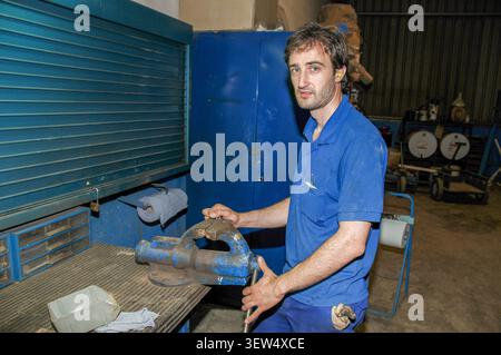 A male industrial worker in a blue uniform holds a heavy metal vise in a workshop. Tools, cabinets, and equipment surround him in a functional mechani Stock Photo