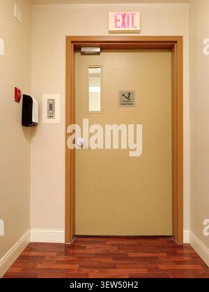 A fire door exit is shown at the end of a hallway. The door has a push bar and clear exit signs above it. Stock Photo