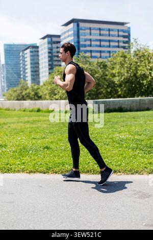 Caucasian athletic man in black sportswear jogs on a park path near modern office buildings, urban fitness and healthy lifestyle concept. Stock Photo
