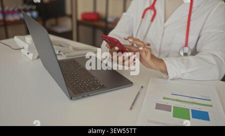 Woman doctor taps red smartphone with hands and stethoscope beside laptop on desk inside building; focus. Stock Photo