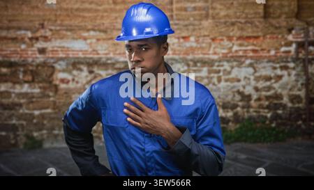 Man construction worker, african american, in blue hardhat and blue coveralls, places hand on chest in front of brick building, serious expression and Stock Photo