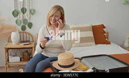 Woman holding creditcard and smartphone to ear while sitting on bed beside open suitcase and straw hat in building; trip planning anticipation. Stock Photo