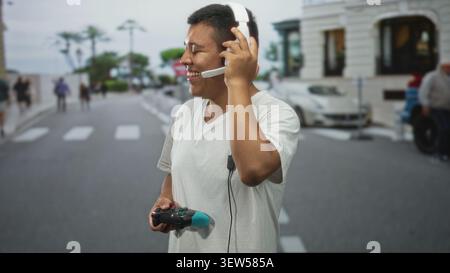 Man holding game controller and touching headset microphone on a city street crosswalk, smiling while playing; joyful play. Stock Photo