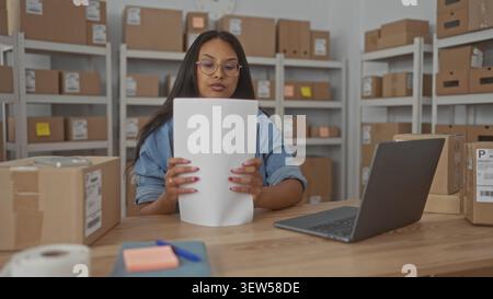 Woman typing on laptop and handling shipping papers at packing desk in warehouse storage room; entrepreneurial focus. Stock Photo